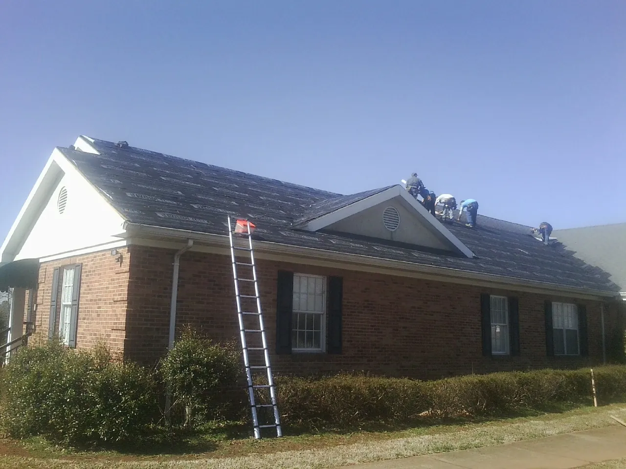 Crew tearing shingles off a brick ranch roof with a ladder set — Emergency Roof Repair work by our crew in Kewaskum