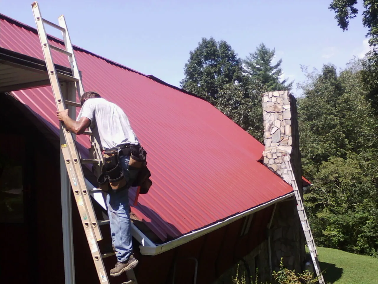 Roofer on a ladder beside a red metal roof and stone chimney — Soffit & Fascia Repair by Roofing Brothers in Kewaskum